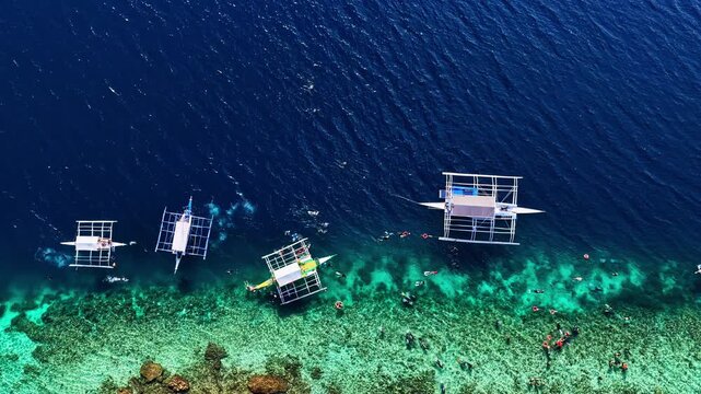 Stunning top-down drone view over Moalboal, Philippines. Features traditional outrigger boats anchored on a dramatic coral reef drop-off filled with snorkelers and divers in clear turquoise water.