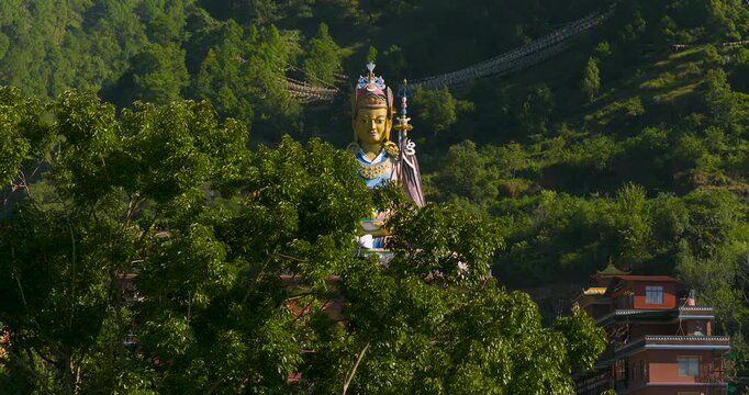 Drone lateral view moving from lush green tree bushes before clearing them to reveal the massive, colorful Guru Rinpoche statue on Pharping, Nepal