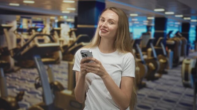 Woman holding smartphone with both hands, smiling and looking up slightly in indoor gym studio filled with cardio machines and equipment; calm focus routine.