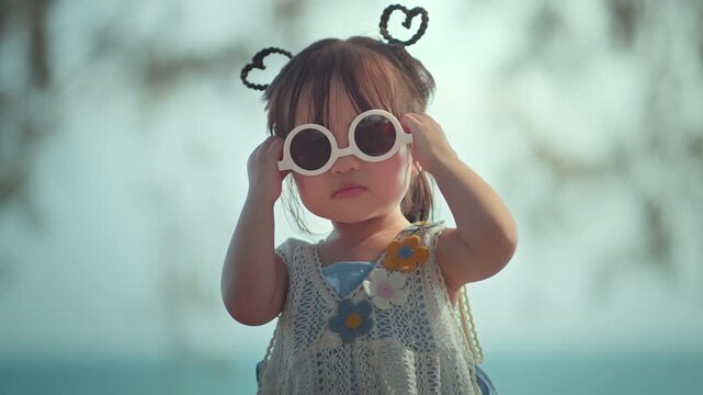 Happy Asian toddler girl smiling and wearing cute white sunglasses outdoor, cheerful little child enjoying summer vacation concept for happy childhood, parenting, and kids fashion lifestyle