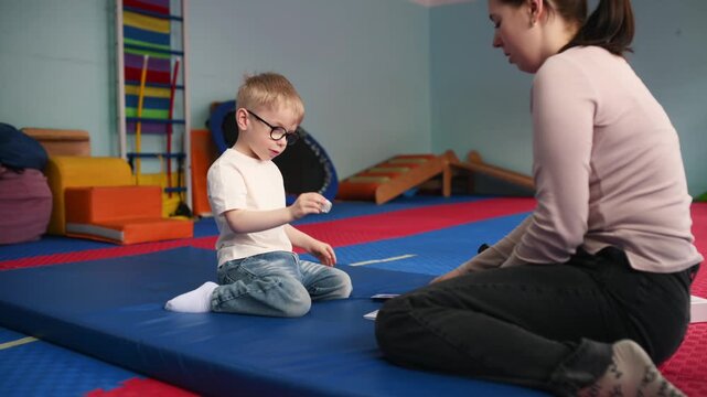 A small child wearing glasses sniffs and guesses smells while playing an educational game. on a blue mat in a bright indoor play space with various soft play structures.