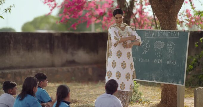 Indian young adult woman do dance teach to girl boy enjoy day under tree rural area teen child group wear uniform sitting on mat look tutor learn basic step moves have fun joy open class field place