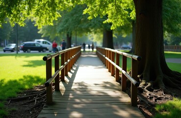 Fototapeta premium Wooden bridge crosses green park lawn with large trees, sunlight filters through leaves. People walk in background on sunny day. Cars drive nearby on street.