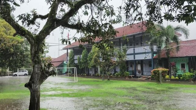 Cinematic wide shot of a traditional Indonesian school building under heavy tropical rain with a large mossy tree and a flooded wet grass field.