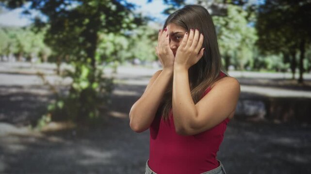 Woman covers face with hands in park, palms pressed to cheeks, sleeveless red top visible, sunlight and trees behind her; anxiety vulnerability.