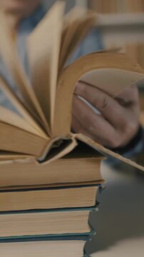 A man is reading a book, examining the pages carefully. Several books are stacked beneath his hands, indicating a moment of focused study or research.