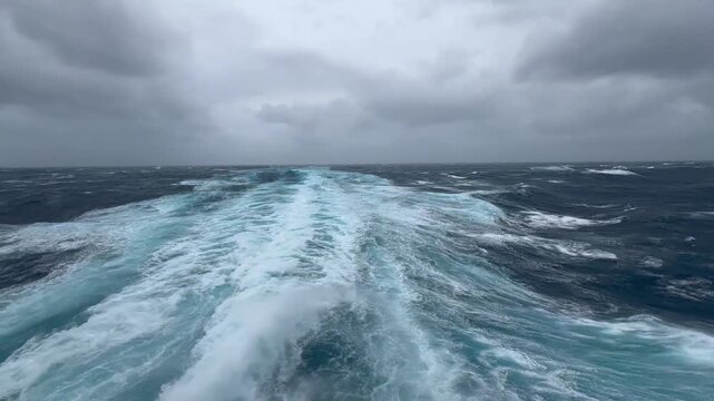 Raw footage from the stern of a cruise ship mid-Atlantic in a severe storm. Heavy rolling, massive waves, dark overcast sky. Two propeller trails visible through a foamy wake battered by wind-torn.