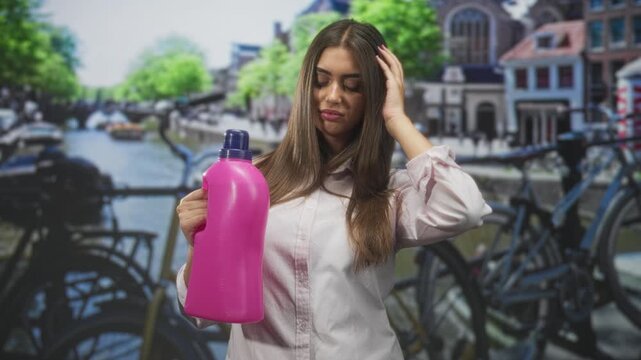 Young hispanic woman holds bright pink detergent bottle, touches hair and looks unsure in studio; skepticism concern.