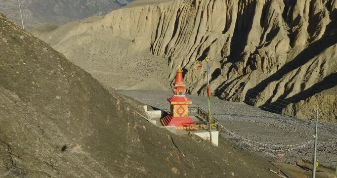 Aerial view captures a vibrant, orange-red Buddhist chorten standing beside a mountain road in Mustang, Nepal, contrasted against stark, wind-eroded cliffs.