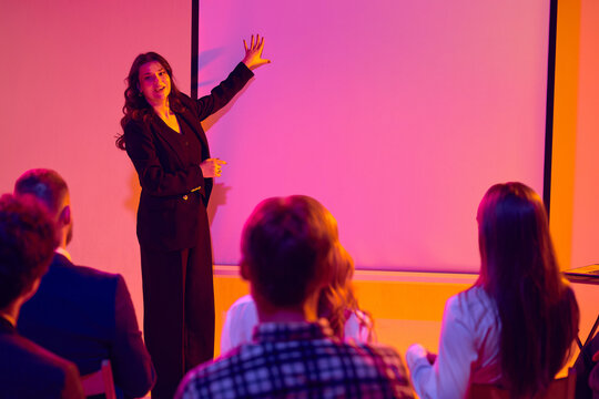 Presenter explaining ideas beside projection screen to seated audience. Concept of corporate presentation, seminar coaching, business education, teamwork and communication skills.