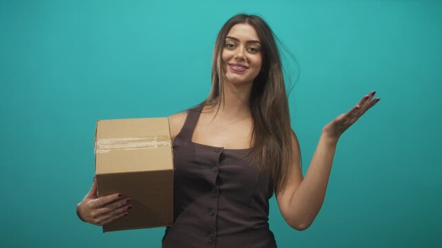 Woman holds cardboard box with left hand and shrugs with right open palm in studio teal backdrop; playful surprise.
