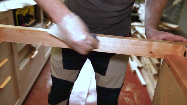 Professional carpenter in protective gloves working in workshop with wooden products, joiner applying gold colour paint on surface of raw wooed plank, worker varnishing wood for further furniture