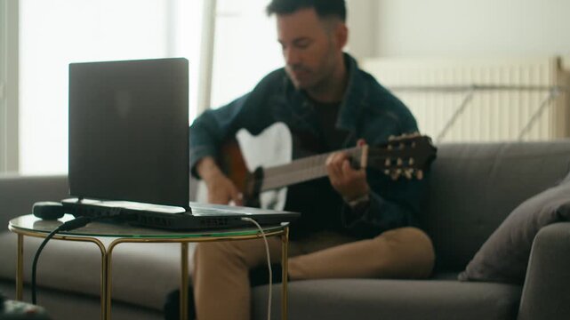 An authentic scene of a young man practicing acoustic guitar on a sofa while following an online lesson on his laptop. Great for remote learning and hobby concepts.