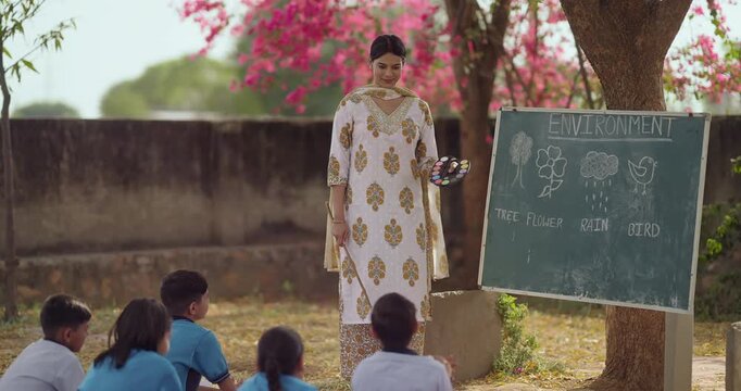 Indian happy young adult female standing hold color palette talk speak to girl boy group under tree rural area field tutor pointing stick on words teach look board read do study learn open class place