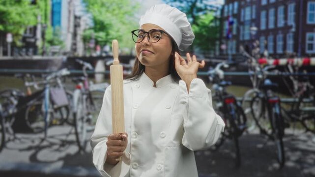 Woman chef wearing toque and white coat holding a rolling pin and making an ok sign with fingers in street; confidence culinary skill.