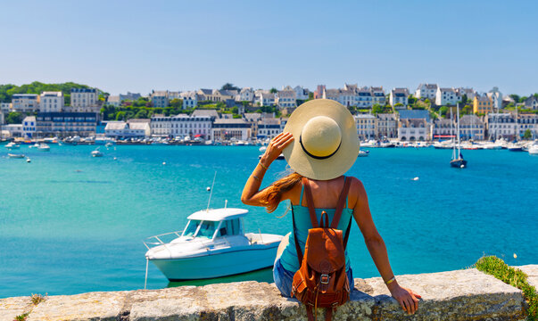 woman tourist with straw hat and backpack looking at coastal town harbor in Brittany in France, authentic summer travel and vacation concept