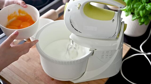 An electric stand mixer whips fluffy white batter in a bowl with a dish of egg yolks beside it in a modern kitchen baking a homemade sponge cake from scratch.