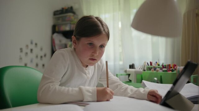 A diligent young girl sits at her desk, writing in a notebook and using a tablet for her homework. She is focused on her studies during an online class.
