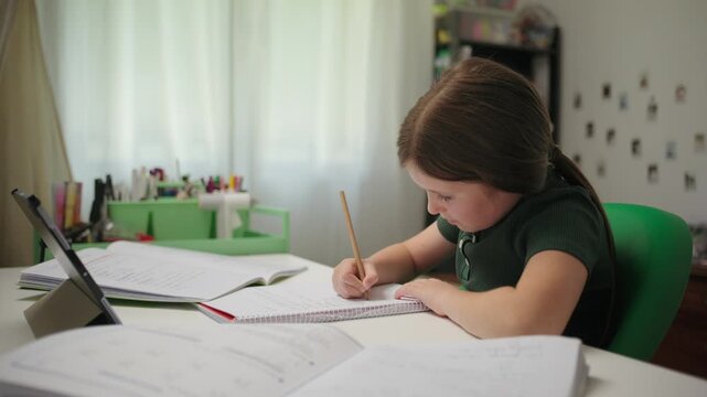 A diligent elementary school girl studies at home, writing in a notebook with a pencil while doing her homework.
