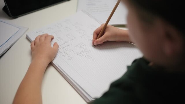 Over the shoulder view of a young student writing with a pencil in a notebook. The child is solving math problems and doing homework at a white desk.