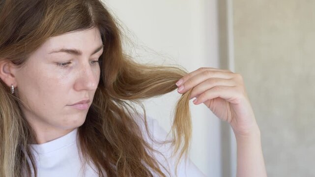 Woman pulls and examines damaged hair strand checking breakage loss and split ends in haircare concern closeup. Footage suits anti-breakage shampoo brand, trichology clinic content, hair loss beauty