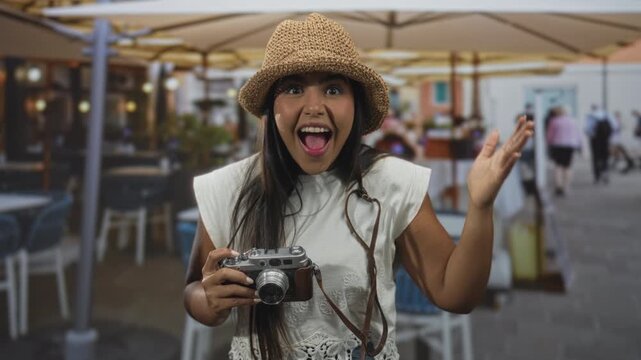 Woman holding vintage film camera shields eyes with hand under straw hat near outdoor cafe tables on busy pedestrian street; travel curiosity surprise.