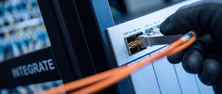 Fiber optic cable network connection inside datacenter server rack with IT technician hand plugging SFP transceiver module into switch port for high speed internet database system integration.