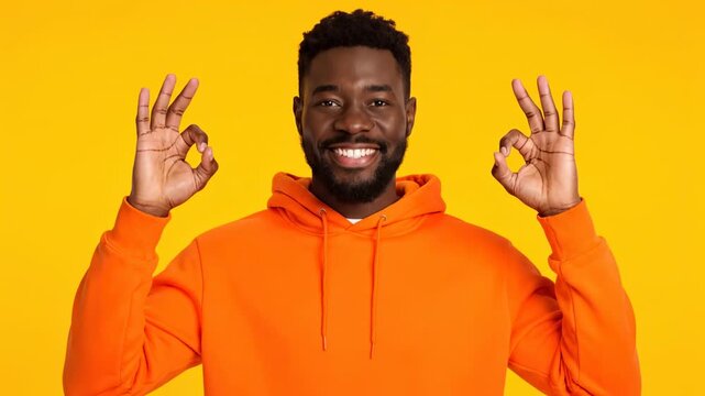 A smiling man in orange hoodie makes an "OK" gesture against a yellow backdrop