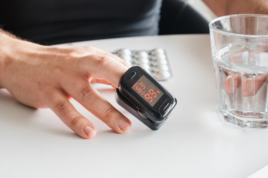 Close-up of a fingertip pulse oximeter showing the oxygen reading on a mature man's finger beside a glass of water and pills illustrating home health monitoring and self care.
