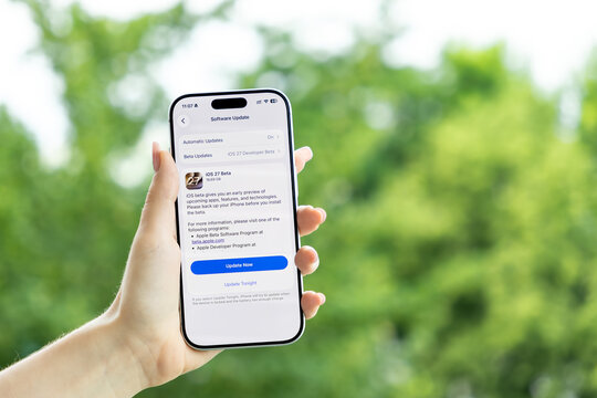 Poznan, Poland - June 9, 2026: Hand holding a smartphone displaying an iOS 27 developer beta update screen on a bright outdoor background, symbolizing mobile software development