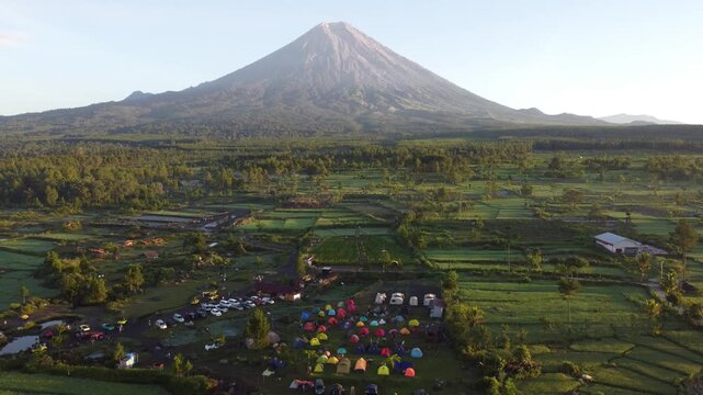 Drone view of Mount Semeru overlooking the agricultural landscape of Pronojiwo, East Java, with green farmland and volcanic scenery illuminated by morning light.