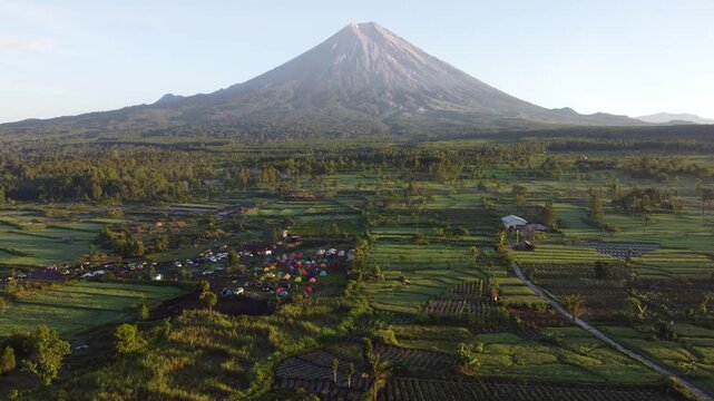 Drone view of Mount Semeru overlooking the agricultural landscape of Pronojiwo, East Java, with green farmland and volcanic scenery illuminated by morning light.