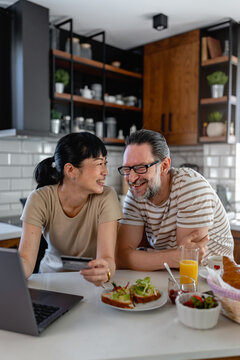 Cheerful couple looking at laptop screen in kitchen during breakfast. Remote work, online communication, technology use and connected home lifestyle