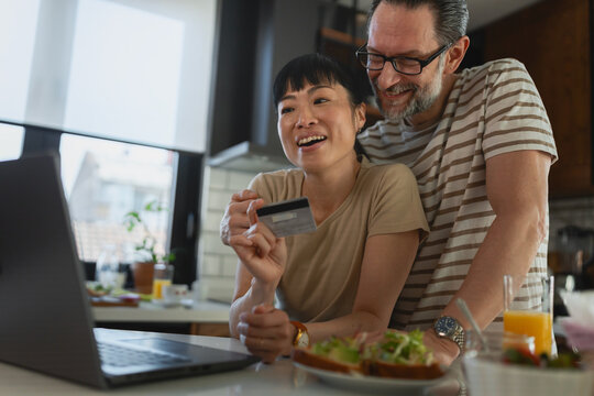 Happy multicultural mature couple shopping online from home, using a laptop and credit card in the kitchen. Lifestyle portrait representing e-commerce, digital banking, and family finances