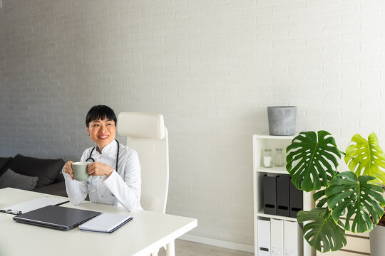 Smiling physician enjoying a coffee break while working in bright clinic. Doctor lifestyle, wellbeing, productivity and modern healthcare concept