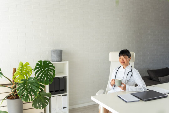Smiling physician enjoying a coffee break while working in bright clinic. Doctor lifestyle, wellbeing, productivity and modern healthcare concept