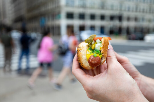 Close-up of hand holding traditional Chicago style hot dog on street city background. Fast food, american cuisine, street food culture and summer lunch concept.