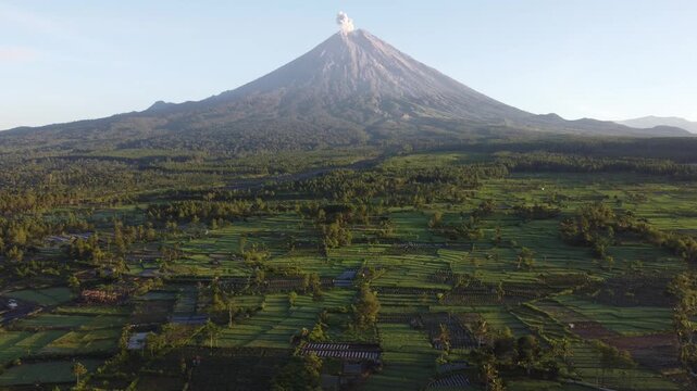 Drone view of Mount Semeru overlooking the agricultural landscape of Pronojiwo, East Java, with green farmland and volcanic scenery illuminated by morning light.