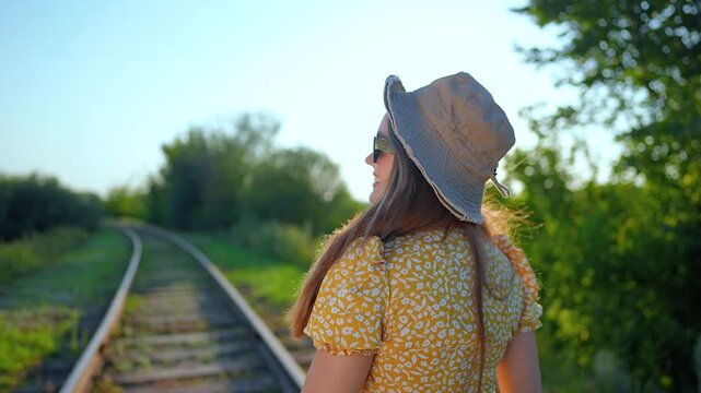 Happy woman in yellow dress is standing on railroad track. Female traveler with hat looks back and smiles outdoors. Girl walks in nature on train tracks. Sun light shines on happy woman in summer.