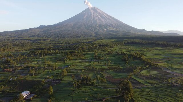 Drone view of Mount Semeru overlooking the agricultural landscape of Pronojiwo, East Java, with green farmland and volcanic scenery illuminated by morning light.