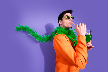 Obraz premium Stylish man in orange suit and green feather boa sipping a green cocktail with sunglasses against a vibrant purple background, playful retro party fashion portrait