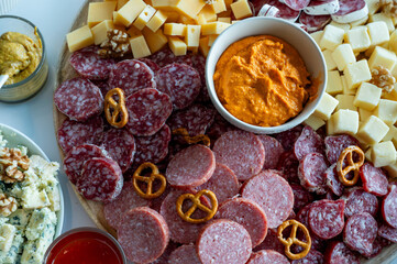 Fototapeta na wymiar A round sharing board with salami slices cheese cubes pretzels walnuts and a red pepper dip on a white table a festive charcuterie platter for entertaining.
