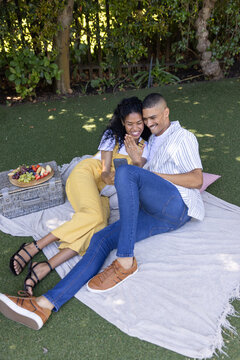 Couple reclining on light gray picnic blanket on green lawn, leaning and looking at smartphone
