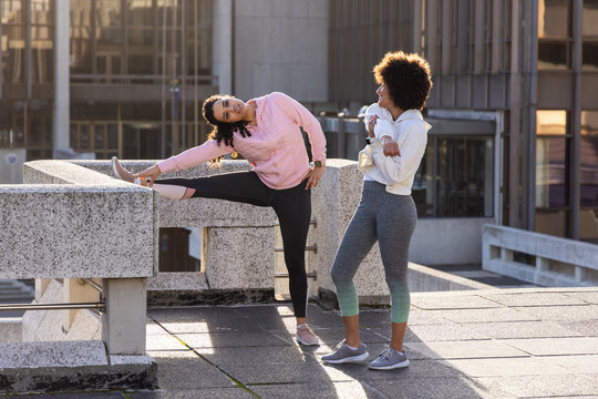 Diverse female friends stretching and chatting in hoodies on rooftop using parapet and sneakers