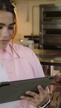 Vertical video: Looking at tablet, woman in pink shirt tapping, scrolling at bakery, checking stock