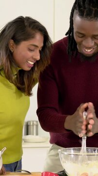 Vertical video: Leaning woman in sweaters encouraging man whisking pale batter at counter with bowl