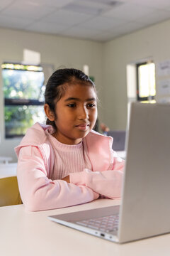 Indian school-age girl sitting at white desk using silver laptop in classroom wearing pink hoodie