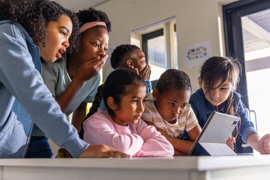 Diverse group of school-age children leaning around tablet in classroom, holding folio and watching