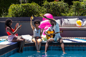 Obraz premium African American friends sitting in swimwear at pool, playing guitar and holding drinks by flamingo