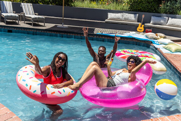African American friends floating on sprinkle donut and pink ring in backyard pool, holding cup © wavebreak3